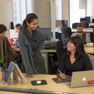 K7316 Hannah Foster Intern for Hello Parent A woman stands to chat with another woman sitting down working at a computer.