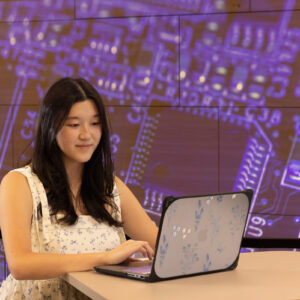 K3825 Serena Owens Computer Graduate Student A young woman works on her laptop in front of a purple background showing a computer motherboard.