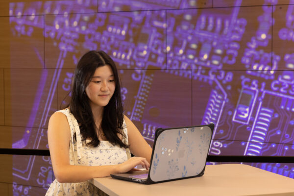 A young woman works on her laptop in front of a purple background showing a computer motherboard.