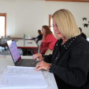 K11226 Debra Saunders Brown news story An older woman works on her laptop while looking at her notes.